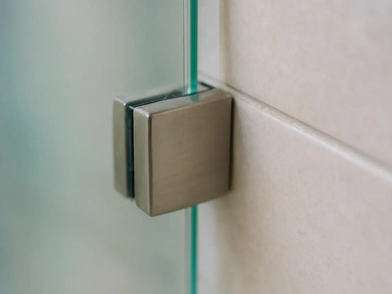 A brushed metal hinge clamps a glass panel to a beige tiled wall