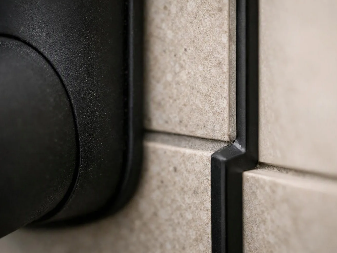 Close-up of a black plastic fixture mounted beside beige textured tiles with a vertical seam