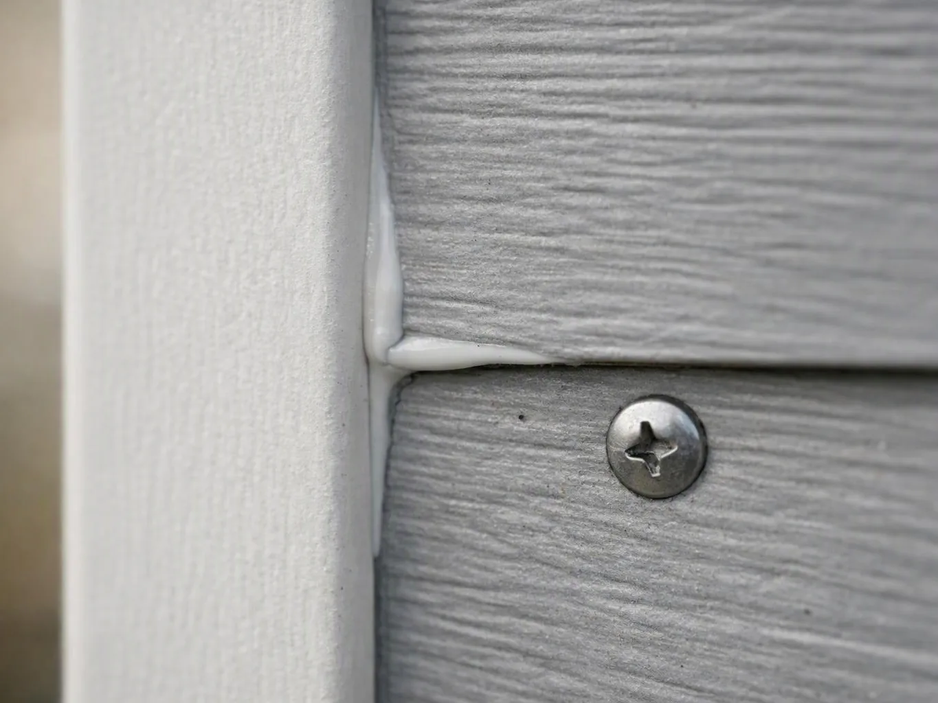 Close-up of gray siding meeting a white trim board, with a visible screw and caulk seam