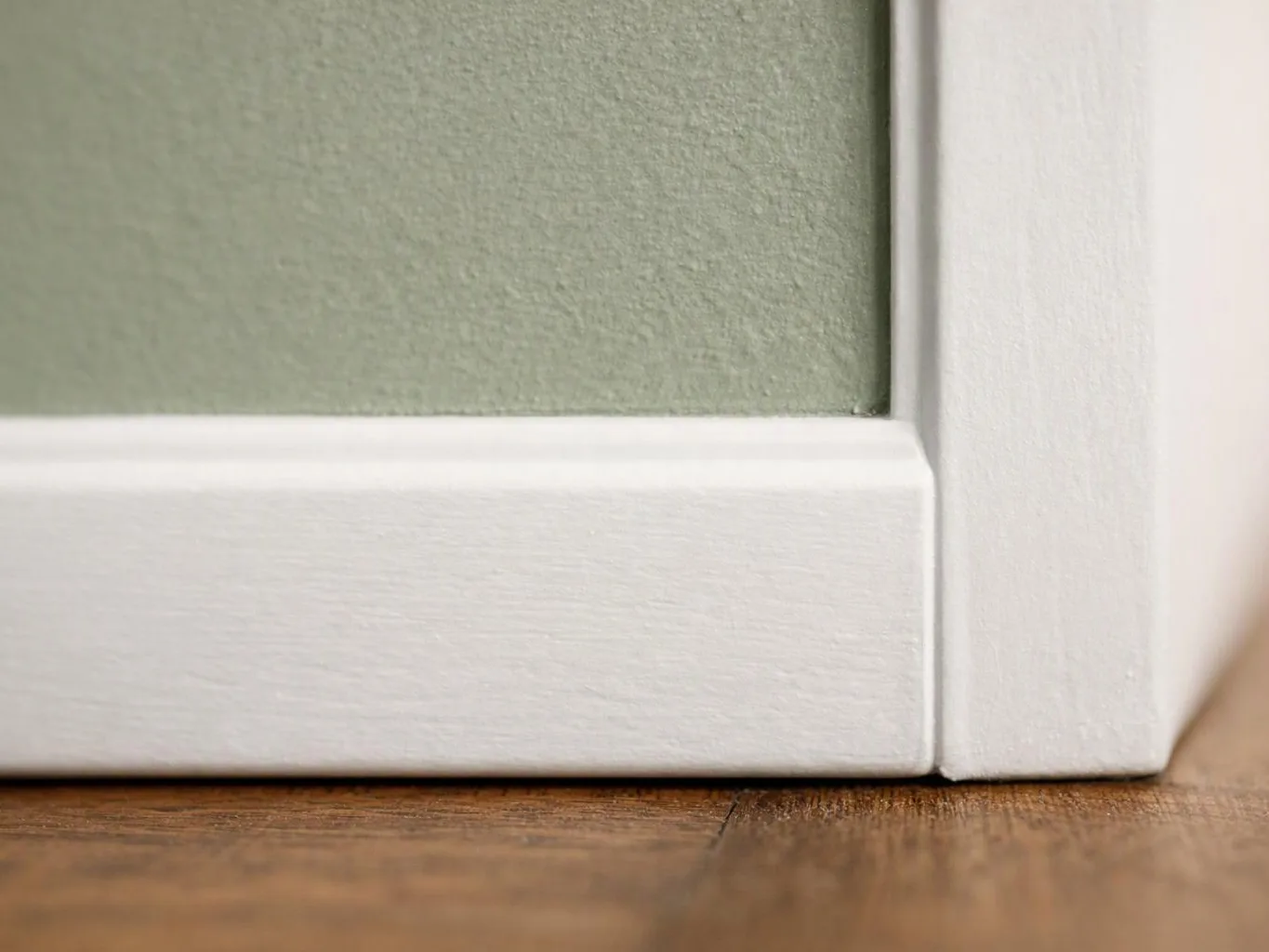 Close-up of a white baseboard and door frame against a pale green wall above a wooden floor